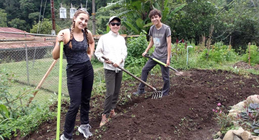 Three people stand in a dirt plot holding gardening tools. They are smiling. 